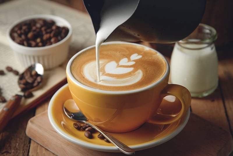 Tách cà phê cappuccino với họa tiết latte art, sữa đang được rót vào từ bình, bên cạnh là hạt cà phê.