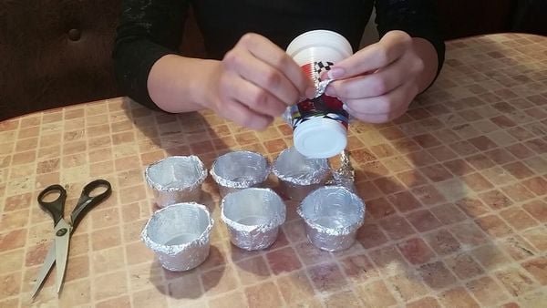 Người đang bọc cốc nhựa bằng giấy bạc tr&ecirc;n b&agrave;n, b&ecirc;n cạnh l&agrave; k&eacute;o v&agrave; s&aacute;u cốc kh&aacute;c đ&atilde; bọc xong.