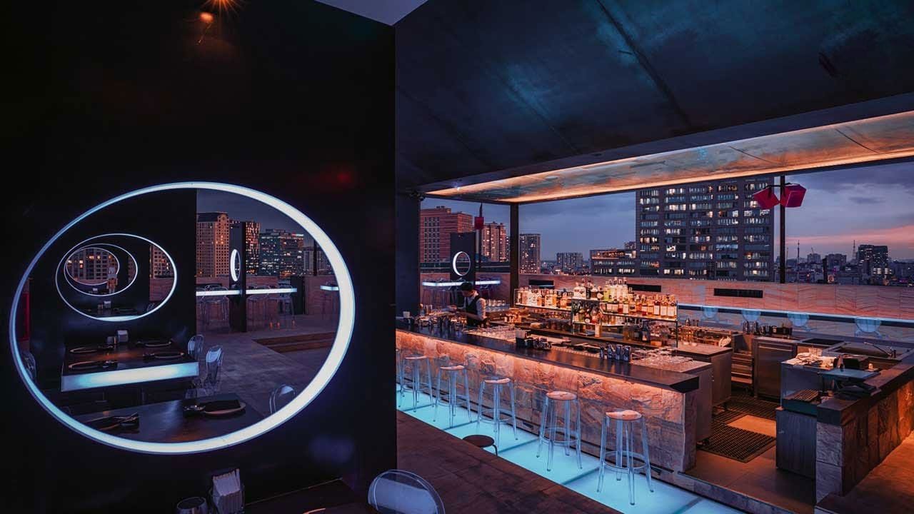 Quầy bar trên sân thượng với ánh sáng neon, bàn ghế hiện đại và khung cảnh thành phố về đêm.