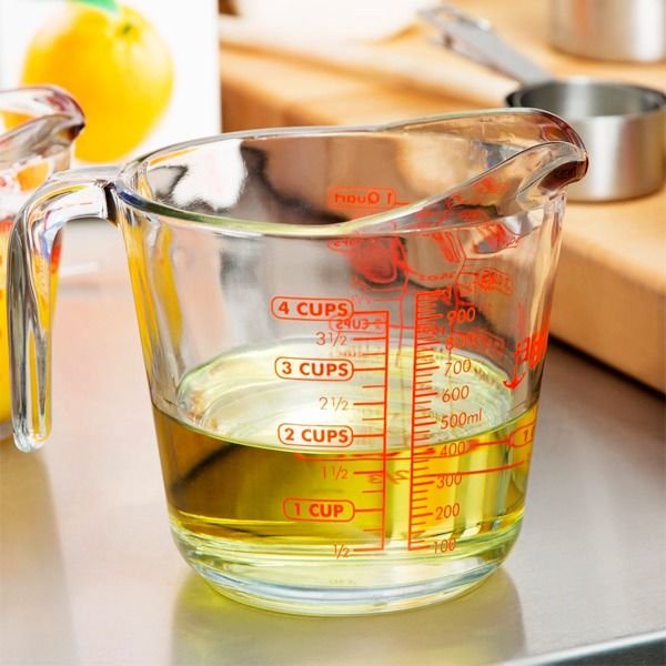 oz-la-gi-1-oz-bang-bao-nhieu-ml_a385145cb9c0488ca8bf4abb5bbbef8f_grande Cốc thủy tinh đo lường có chất lỏng màu vàng bên trong, đặt trên bề mặt bếp với các dụng cụ nấu ăn khác.