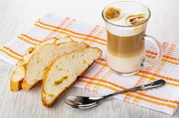 Ly cà phê bên cạnh ba lát bánh mì trên khăn trải bàn sọc cam, có một chiếc thìa kim loại.