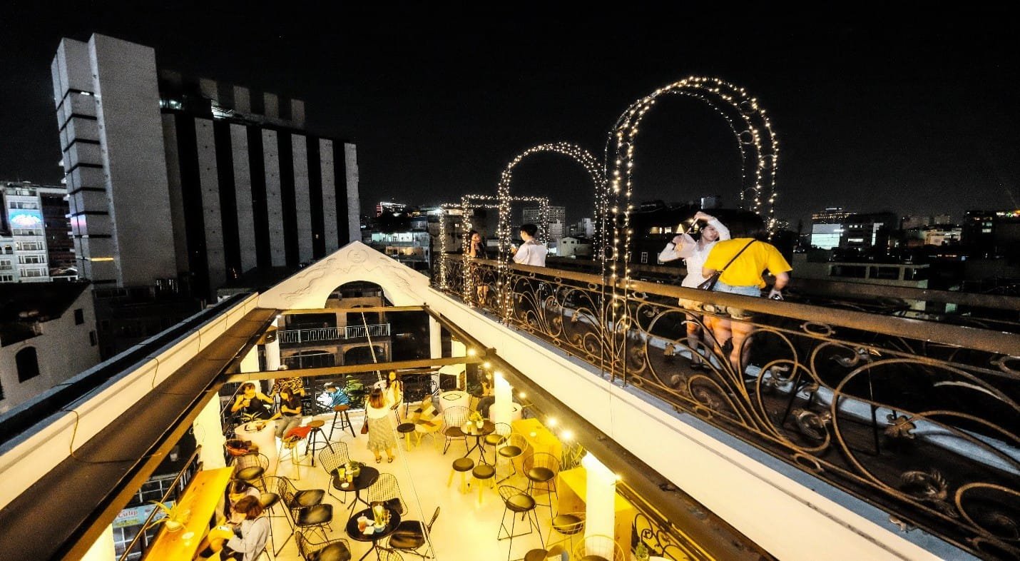 Quán cà phê trên sân thượng vào ban đêm, trang trí đèn và có người chụp ảnh, nhìn ra tòa nhà cao tầng.