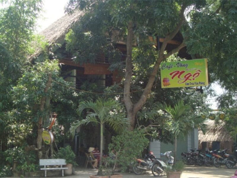 Gợi ý 14 quán cà phê chòi mát mẻ, thư giãn tại TPHCM 12 Quán cà phê với mái tranh, biển hiệu màu vàng, xung quanh có nhiều cây xanh và xe máy đậu phía trước.
