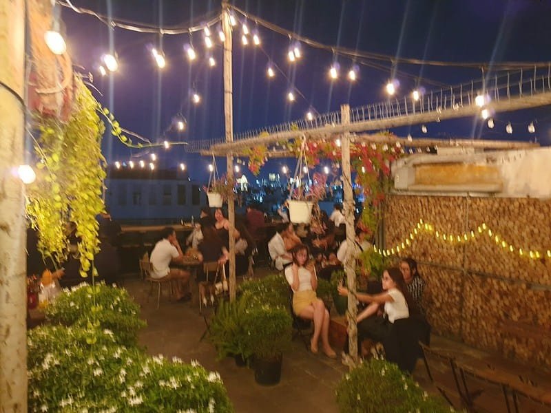 Quán cà phê ngoài trời vào ban đêm với nhiều người ngồi dưới ánh đèn dây và cây xanh trang trí.