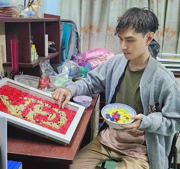 Người đang sắp xếp hạt nhựa màu trên khung tranh chữ, tay cầm tô chứa hạt nhựa, ngồi cạnh bàn làm việc.