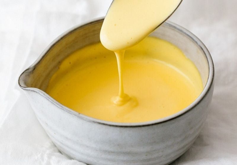 Bát sứ đựng sốt màu vàng, một chiếc thìa đang múc sốt chảy xuống bề mặt.