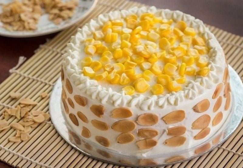 Chiếc bánh kem trang trí hạt bắp vàng và lát hạnh nhân, đặt trên bàn trải chiếu tre.