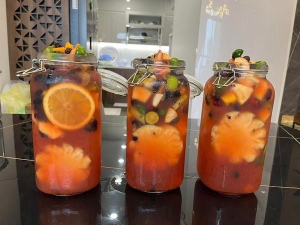 tra-trai-cay-nhiet-doi_9ef3970c9f9a4cfcade5ec96f6cc7a35_grande Ba lọ nước trái cây nhiều màu sắc với lát cam và dứa bên trong, đặt trên mặt bàn bếp.