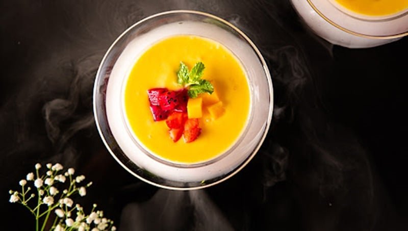 Ly tr&aacute;ng miệng m&agrave;u v&agrave;ng, trang tr&iacute; với tr&aacute;i c&acirc;y tươi v&agrave; l&aacute; bạc h&agrave;, đặt tr&ecirc;n nền tối cạnh hoa trắng.