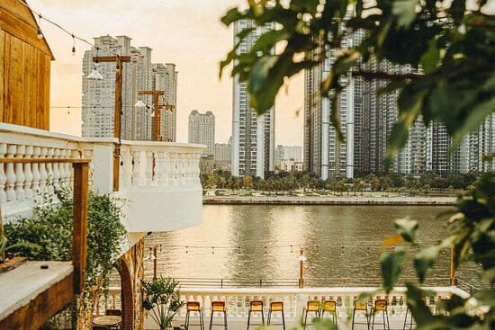 Khung cảnh bờ sông với tòa nhà cao tầng và hàng cây xanh mát, ánh sáng hoàng hôn phủ lên mặt nước.