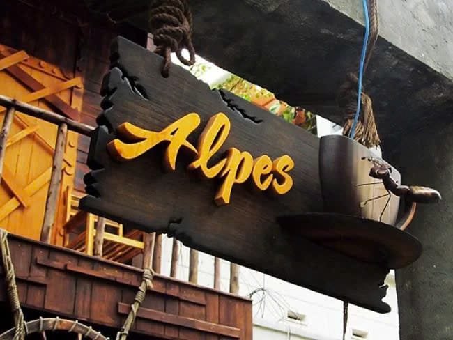 Biển hiệu gỗ với chữ "Alpes" màu vàng, trang trí bằng hình tách cà phê bên cạnh.