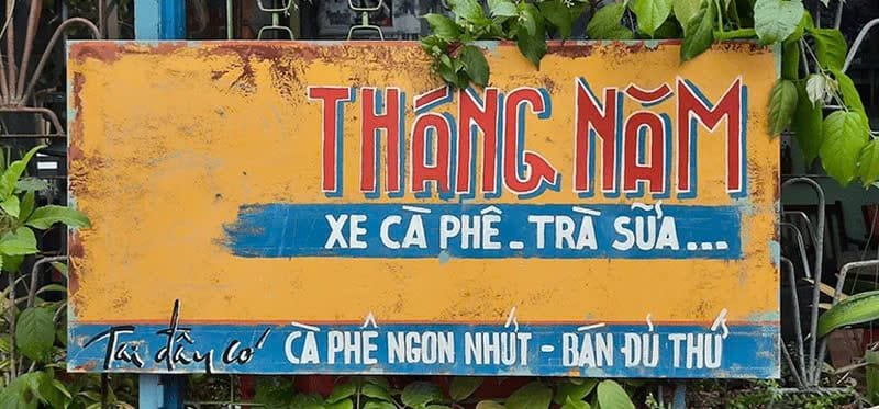 Biển hiệu quán cà phê màu vàng, chữ đỏ và xanh, có cây xanh xung quanh. Nội dung quảng cáo cà phê và trà sữa.