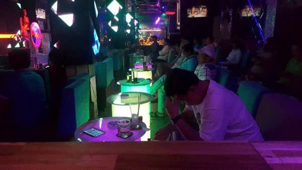Quán bar với ánh sáng neon, bàn ghế, điện thoại trên bàn, nhiều người ngồi thư giãn trong không gian tối.