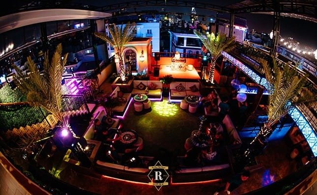 Quán bar trên sân thượng với ánh sáng nhiều màu, cây cọ và nhiều bàn ghế, nhìn ra cảnh thành phố ban đêm.