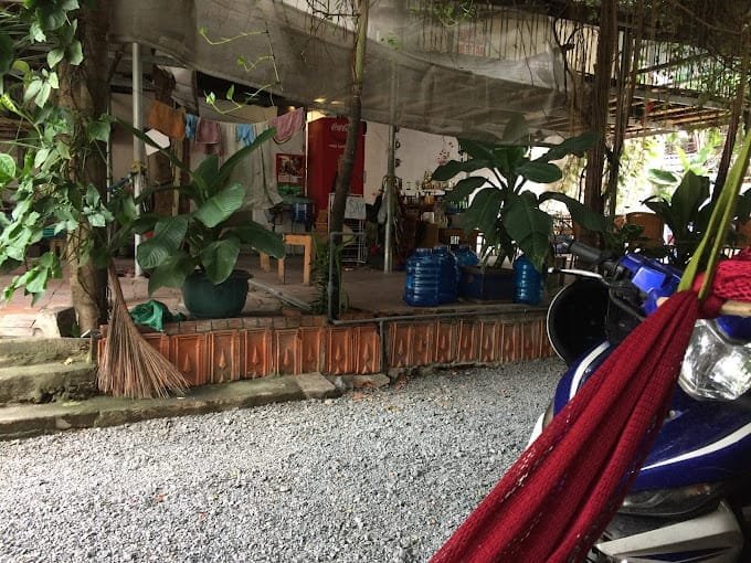 Khu vực ngo&agrave;i trời với ghế, c&acirc;y cảnh, xe m&aacute;y v&agrave; vật dụng treo dưới m&aacute;i che, tạo cảm gi&aacute;c thư gi&atilde;n v&agrave; gần gũi.