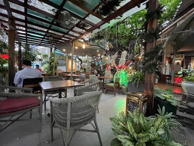 Qu&aacute;n c&agrave; ph&ecirc; ngo&agrave;i trời với b&agrave;n ghế m&acirc;y, c&acirc;y xanh xung quanh, trang tr&iacute; đ&egrave;n v&agrave; c&acirc;y cảnh tạo kh&ocirc;ng gian tho&aacute;ng m&aacute;t.