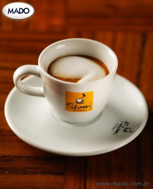 Tách cà phê với lớp bọt sữa trên mặt, đặt trên đĩa trắng, nền gỗ nâu. Logo và chữ trên tách và đĩa.