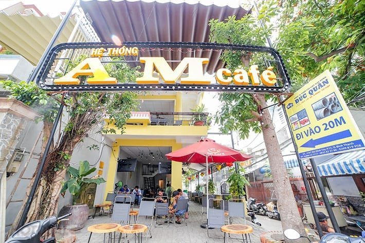 Top 32 thương hiệu nhượng quyền cafe siêu lợi nhuận 18 Quán cà phê ngoài trời với bảng hiệu "AM cafe", có khách ngồi dưới ô dù đỏ và xe máy đậu bên cạnh.