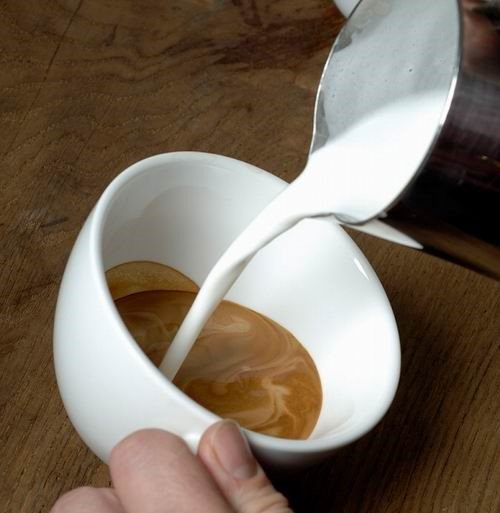 Tay rót sữa vào tách cà phê trắng, tạo hoa văn trên bề mặt, đặt trên bàn gỗ.