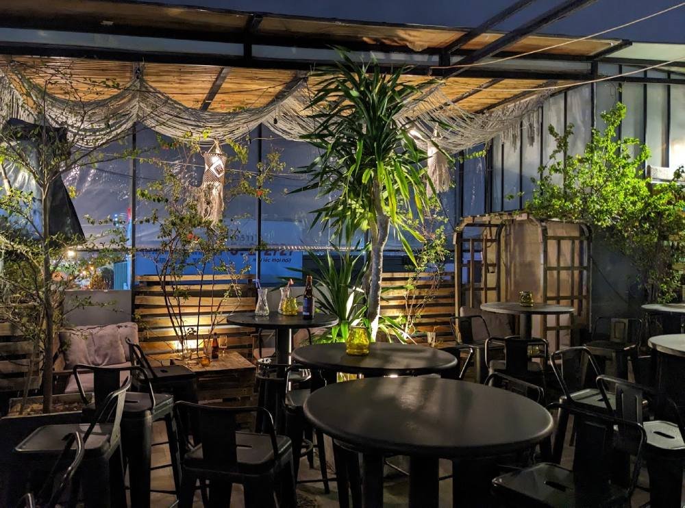 3d5a3364-cafe-da-nang-23 Khu vực quán cà phê ngoài trời vào ban đêm, với bàn ghế và cây xanh trang trí dưới ánh sáng ấm áp.