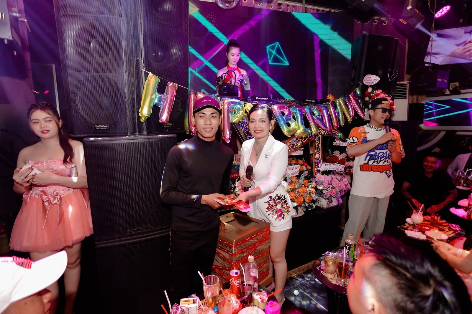 Bữa tiệc sinh nhật với người tặng quà, trang trí bóng bay chữ "HAPPY BIRTHDAY" và hoa tươi.