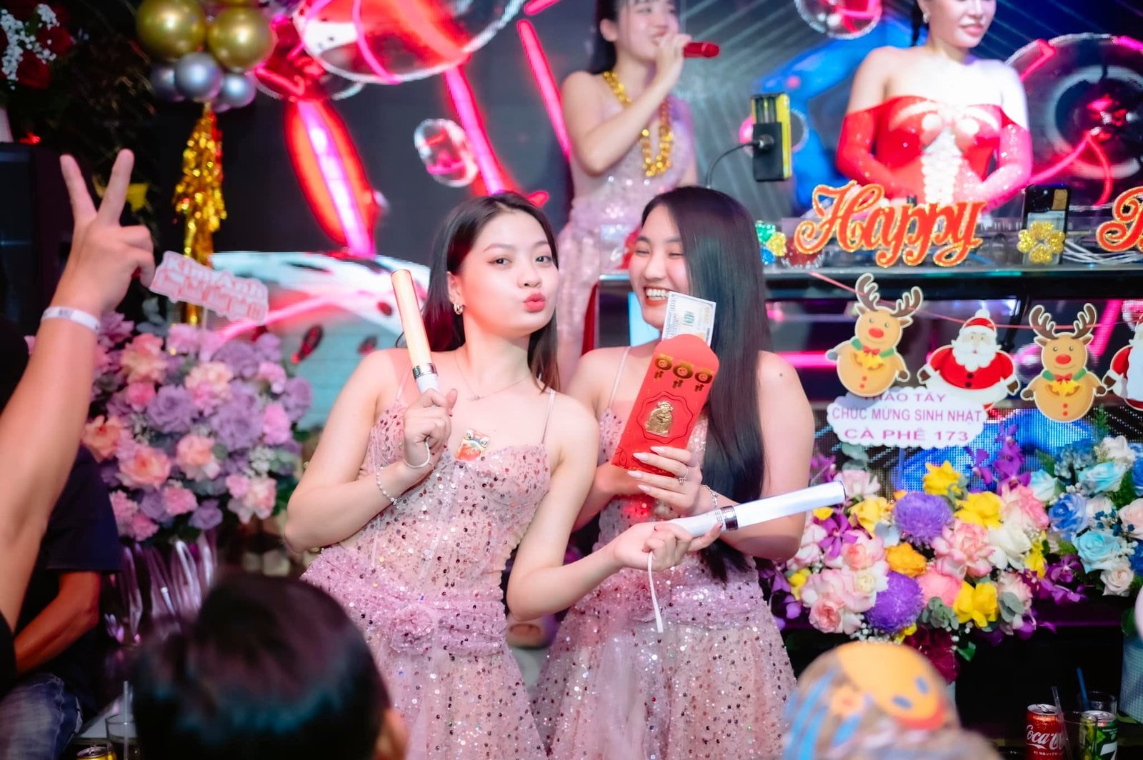 Hai người mặc váy lấp lánh cầm quà và cười trong bữa tiệc sinh nhật với phông nền trang trí màu sắc.