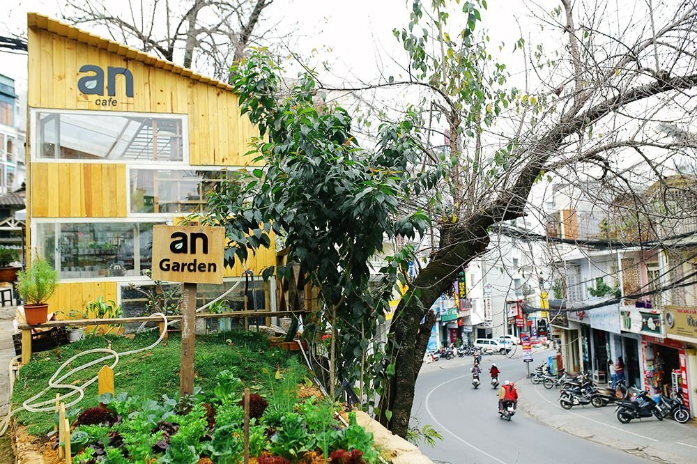 an-coffee-chon-binh-yen-xanh-tham-cho-nhung-tam-hon-mong-manh-26-1635285499 Quán cà phê bằng gỗ với vườn cây phía trước, bên cạnh là đường phố với xe máy đang di chuyển.