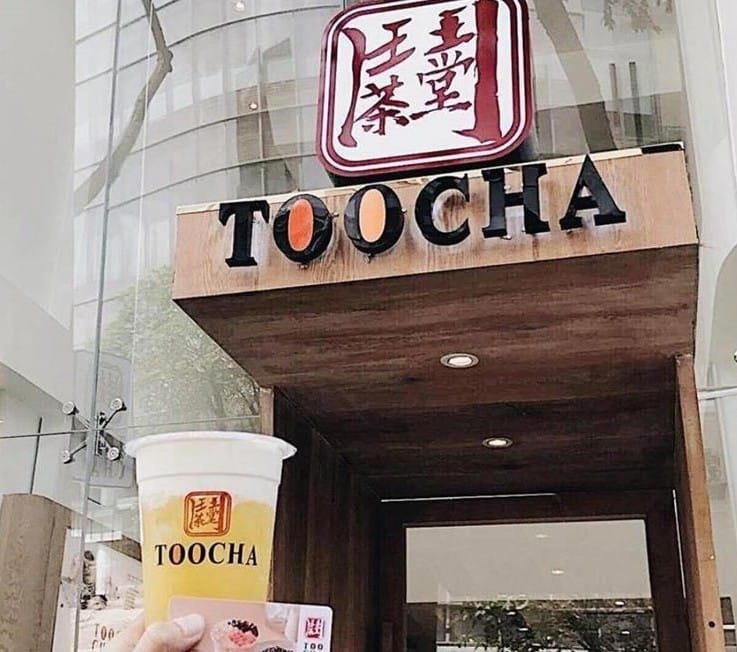 Cốc trà sữa cầm tay trước cửa hàng có biển hiệu "TOOCHA" với thiết kế gỗ và kính hiện đại.