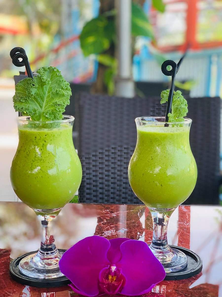 Hai ly sinh tố xanh với rau trang tr&iacute;, đặt tr&ecirc;n b&agrave;n cạnh hoa lan t&iacute;m.
