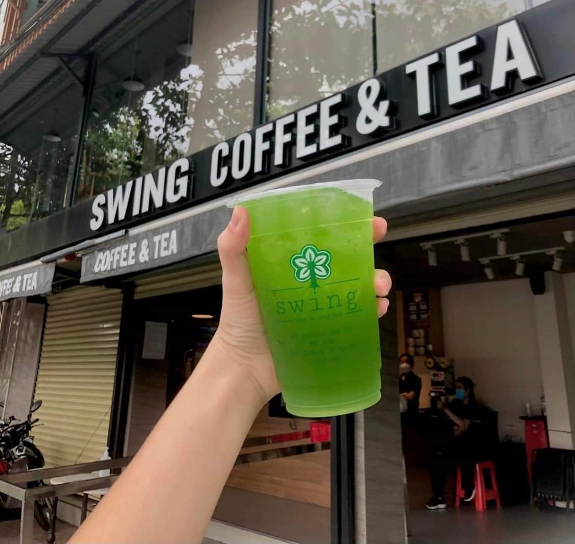 Cốc nước m&agrave;u xanh được giơ l&ecirc;n trước cửa h&agrave;ng c&agrave; ph&ecirc; v&agrave; tr&agrave; c&oacute; bảng hiệu "SWING COFFEE & TEA".