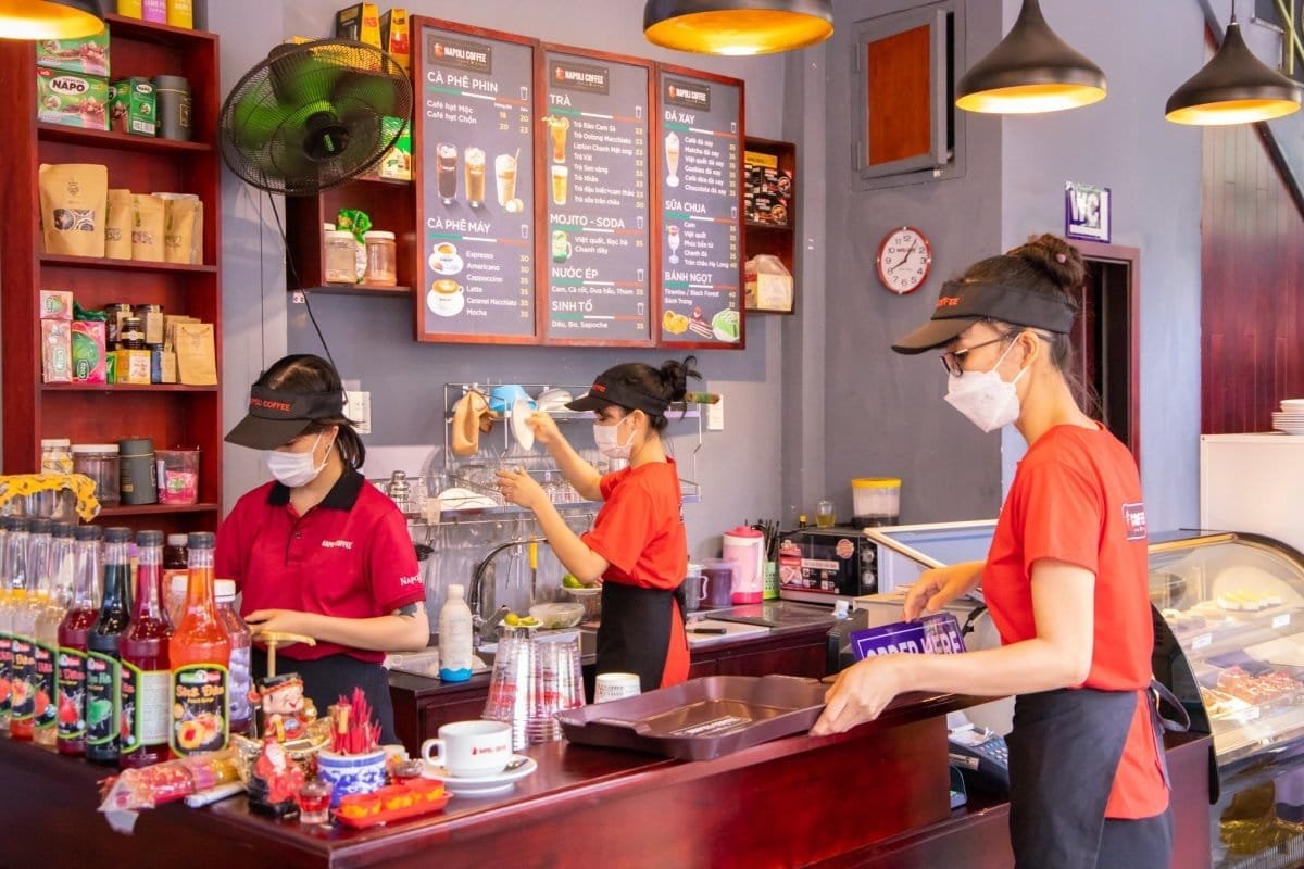 Top 32 thương hiệu nhượng quyền cafe siêu lợi nhuận 12 Nhân viên quán cà phê đang làm việc sau quầy với các loại đồ uống và thực đơn treo trên tường.