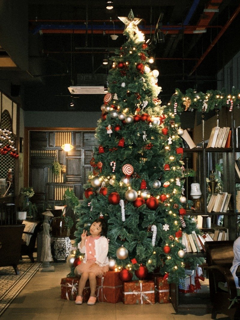 C&acirc;y th&ocirc;ng Noel trang tr&iacute; lộng lẫy với đ&egrave;n v&agrave; quả ch&acirc;u, b&ecirc;n dưới c&oacute; một em b&eacute; ngồi tr&ecirc;n hộp qu&agrave;.