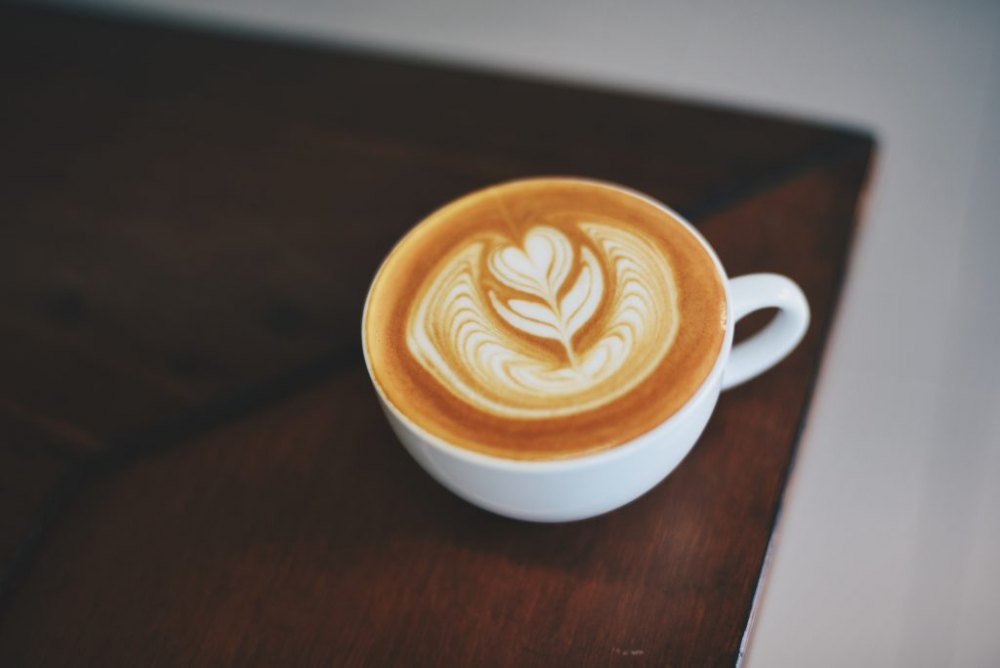 ca-phe-latte Tách cà phê latte trên bàn gỗ, với bọt sữa tạo hình nghệ thuật hoa lá.