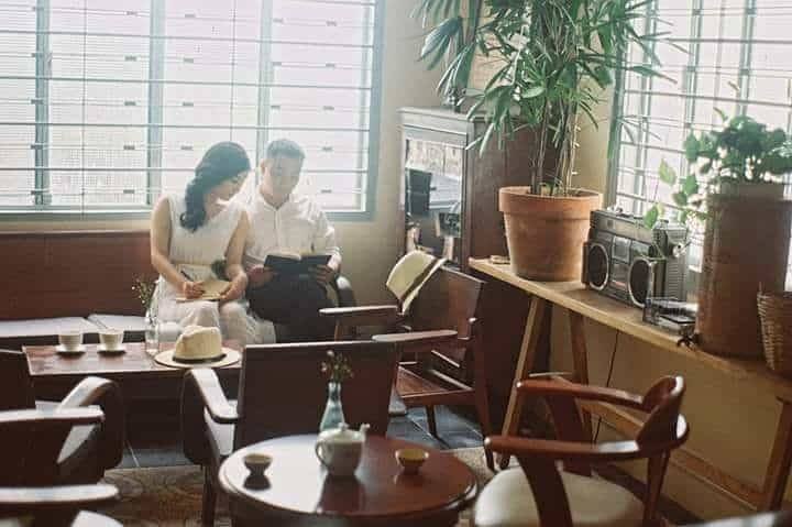 Hai người ngồi trong qu&aacute;n c&agrave; ph&ecirc; cổ điển, đọc s&aacute;ch. Kh&ocirc;ng gian ấm c&uacute;ng với c&acirc;y xanh v&agrave; đồ trang tr&iacute; vintage.