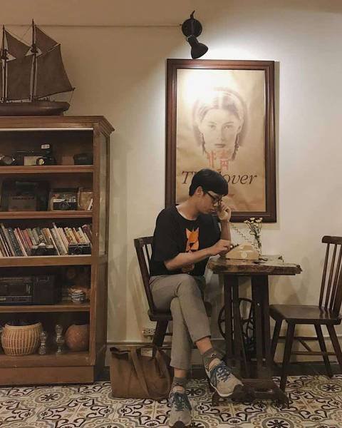 Người ngồi trong qu&aacute;n c&agrave; ph&ecirc;, đang sử dụng điện thoại b&agrave;n. B&ecirc;n cạnh l&agrave; kệ s&aacute;ch v&agrave; tranh treo tường.