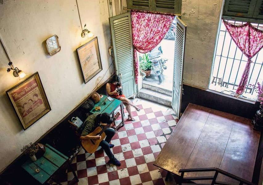 Qu&aacute;n c&agrave; ph&ecirc; với nền gạch đỏ trắng, hai người ngồi cạnh cửa sổ mở, một người chơi guitar.