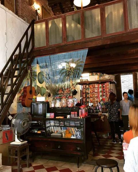 Cửa h&agrave;ng nhỏ với cầu thang gỗ, b&agrave;n quầy h&agrave;ng h&oacute;a, c&acirc;y đ&agrave;n guitar treo tr&ecirc;n tường, v&agrave; v&agrave;i người đứng gần cửa.