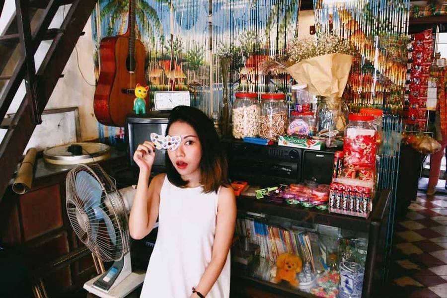 Người phụ nữ trong cửa h&agrave;ng nhỏ, cầm đồ vật che một mắt. Xung quanh c&oacute; đồ ăn nhẹ v&agrave; vật dụng trang tr&iacute;.