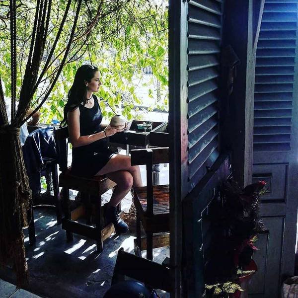 Người phụ nữ ngồi trong qu&aacute;n c&agrave; ph&ecirc;, cầm tr&aacute;i dừa, nh&igrave;n ra ngo&agrave;i cửa sổ, &aacute;nh s&aacute;ng chiếu qua l&aacute; c&acirc;y.