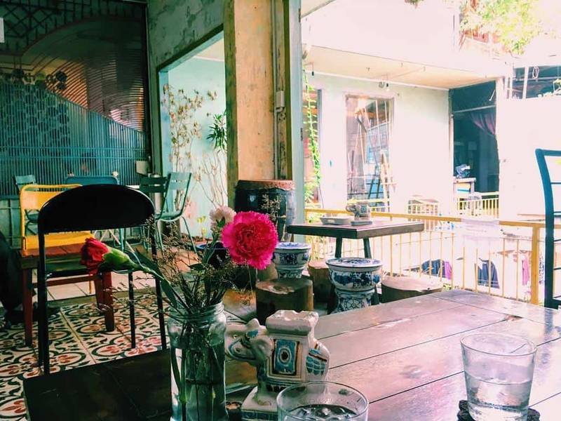 Qu&aacute;n c&agrave; ph&ecirc; phong c&aacute;ch cổ điển với b&agrave;n gỗ, hoa trang tr&iacute; v&agrave; &aacute;nh s&aacute;ng tự nhi&ecirc;n từ cửa sổ.