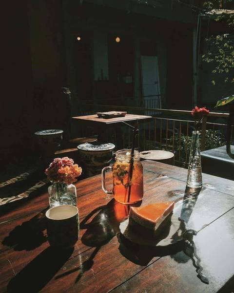 Cốc tr&agrave; đ&aacute; v&agrave; miếng b&aacute;nh tr&ecirc;n b&agrave;n gỗ, b&ecirc;n cạnh l&agrave; lọ hoa nhỏ, &aacute;nh s&aacute;ng tự nhi&ecirc;n chiếu v&agrave;o từ cửa sổ.