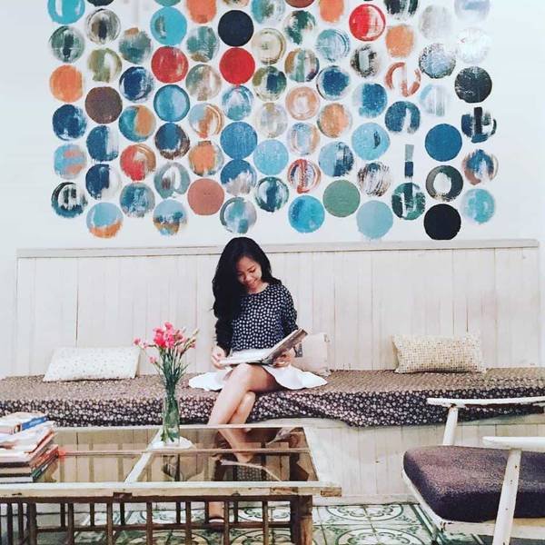 Người đang ngồi đọc s&aacute;ch tr&ecirc;n ghế d&agrave;i, ph&iacute;a sau l&agrave; bức tường trang tr&iacute; với nhiều h&igrave;nh tr&ograve;n m&agrave;u sắc.