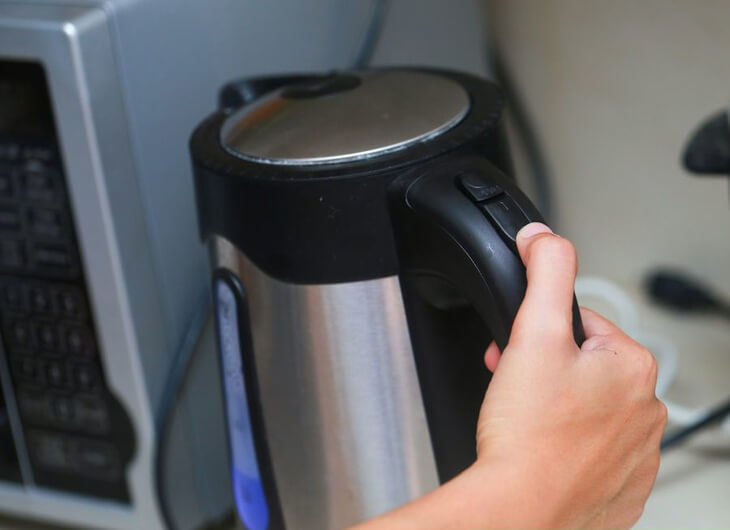 Tay đang cầm ấm đun nước điện, chuẩn bị bật c&ocirc;ng tắc. Ấm đặt gần l&ograve; vi s&oacute;ng v&agrave; ổ cắm điện.