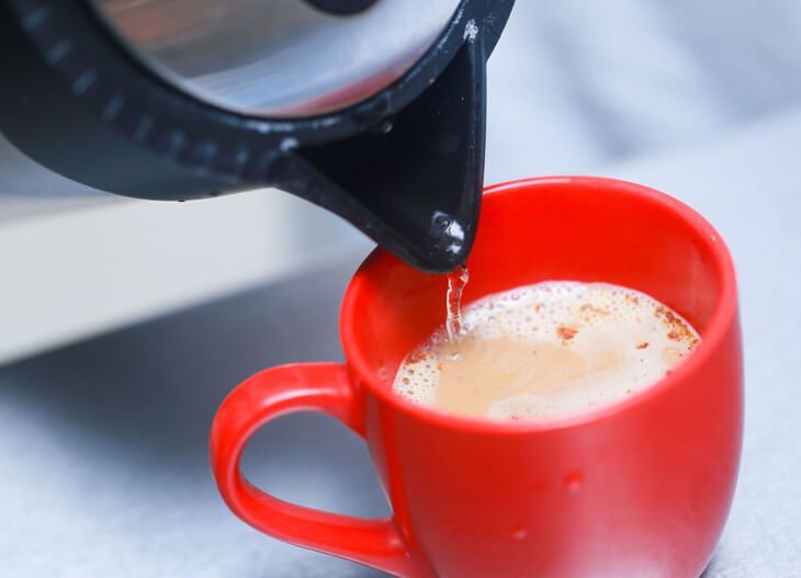 Ấm đun nước đang r&oacute;t nước s&ocirc;i v&agrave;o cốc m&agrave;u đỏ tr&ecirc;n bề mặt m&agrave;u x&aacute;m.