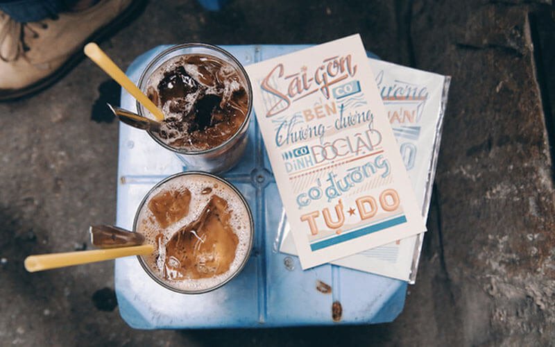 Hai ly cà phê đá trên bàn xanh với tờ giấy in chữ nghệ thuật bên cạnh.