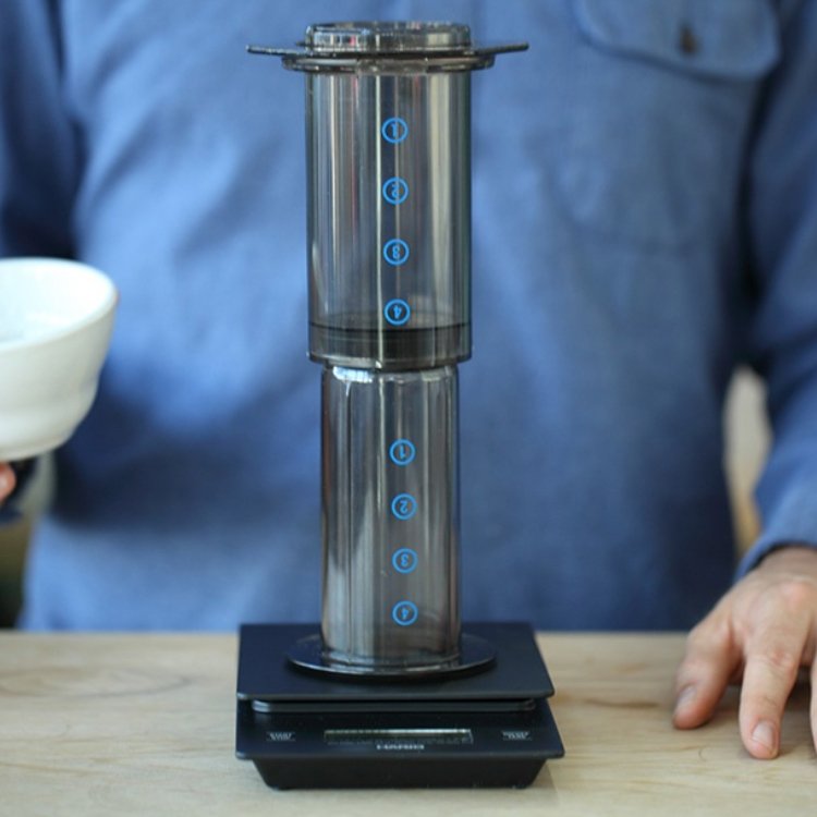 Người mặc áo xanh đang sử dụng dụng cụ pha cà phê Aeropress trên cân điện tử đặt trên bàn.