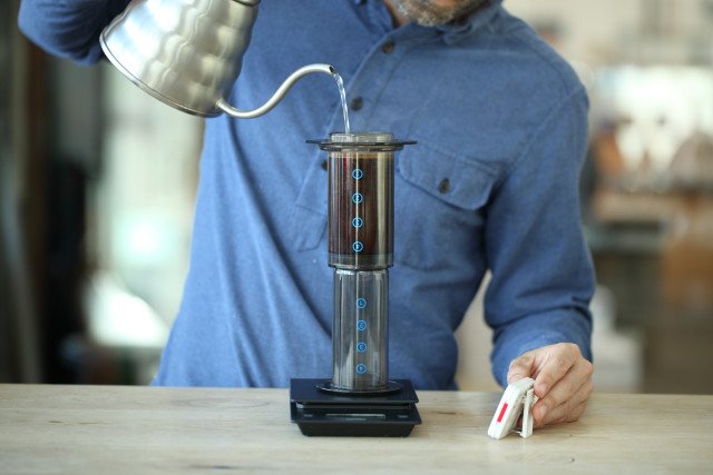 Người đang pha cà phê bằng dụng cụ AeroPress, với nước được rót từ ấm đun, trên cân điện tử.