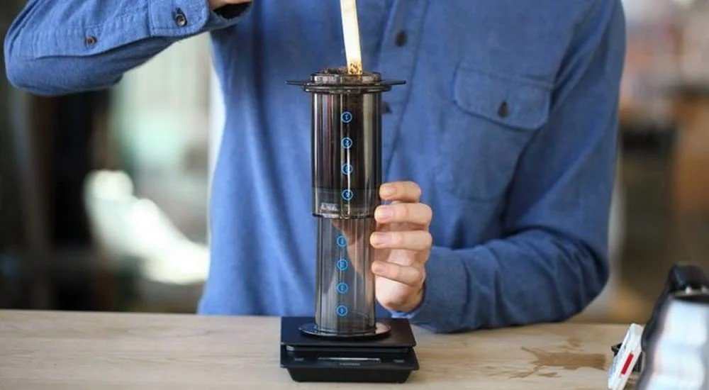 Người đang pha cà phê bằng dụng cụ Aeropress trên bàn gỗ, mặc áo sơ mi xanh.