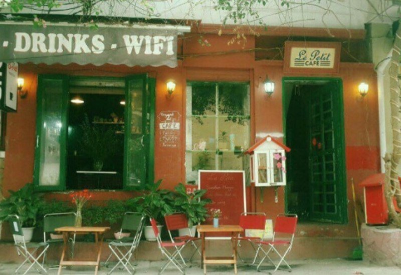 Qu&aacute;n c&agrave; ph&ecirc; nhỏ với cửa sổ xanh, bảng hiệu "Drinks - Wifi" v&agrave; b&agrave;n ghế ngo&agrave;i trời, c&acirc;y xanh xung quanh.