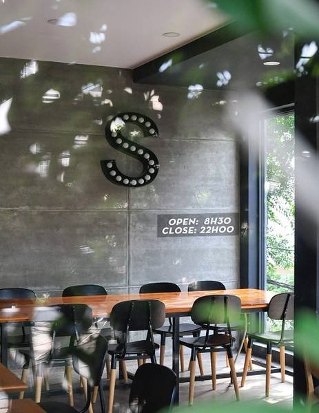 Qu&aacute;n c&agrave; ph&ecirc; với b&agrave;n ghế gỗ, &aacute;nh s&aacute;ng tự nhi&ecirc;n, tường x&aacute;m c&oacute; chữ "S" v&agrave; giờ mở cửa từ 8h30 đến 22h.